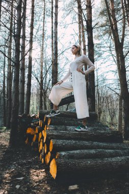 Uzun saçlı sarışın, doğranmış ahşap kütüklerden yapılmış duvarda duruyor, beyaz keten bir kazak giyiyor, geniş pantolonlu, saç bandı takıyor. Ağaçların arasından parlak güneş ışığı