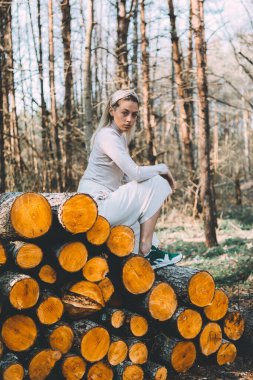 Uzun saçlı sarışın, doğranmış ahşap kütüklerden yapılmış duvarda duruyor, beyaz keten bir kazak giyiyor, geniş pantolonlu, saç bandı takıyor. Ağaçların arasından parlak güneş ışığı