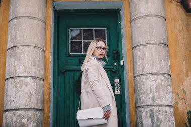 Uzun saçlı, kumlu bir ceket giyen, yün kazaklı, siyah kotlu, eski moda evin arkasındaki merdivenlerde duran sarışın kadın. Mistik bir yer. Pastel renkler.