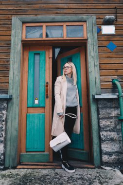 Uzun saçlı, kumlu bir ceket giyen, yün kazaklı, siyah kotlu, eski moda evin arkasındaki merdivenlerde duran sarışın kadın. Mistik bir yer. Pastel renkler.