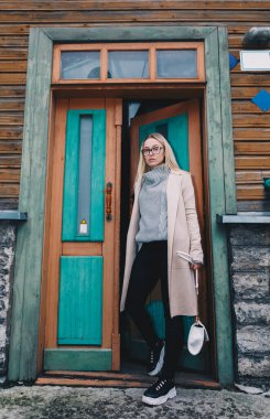 Uzun saçlı, kumlu bir ceket giyen, yün kazaklı, siyah kotlu, eski moda evin arkasındaki merdivenlerde duran sarışın kadın. Mistik bir yer. Pastel renkler.