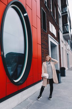 Uzun saçlı, sarı saçlı, yün kazaklı ve siyah kotlu bir kadın, yuvarlak pencereli, modern bir caddede yürüyor. Telefonda konuşuyorlar.