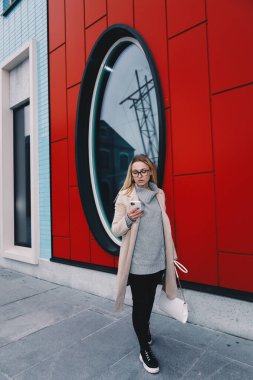 Uzun saçlı, sarı saçlı, yün kazaklı ve siyah kotlu bir kadın, yuvarlak pencereli, modern bir caddede yürüyor. Telefonda konuşuyorlar.