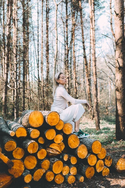 Long haired blonde, standing on the wall of chopped wooden logs, wearing a white linen sweater, white wide pants, headband. Bright sunlight through trees