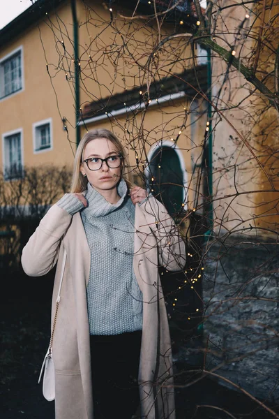 Portrait of long haired blonde woman wearing a sand coat with wool sweater, and black jeans, standing behind old fashioned house. Mystical place with light garland.