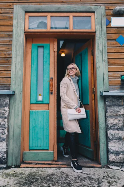 Long haired blonde woman wearing a sand coat with wool sweater, and black jeans, standing on a stairway behind old fashioned house. Mystical place. Pastel colors.