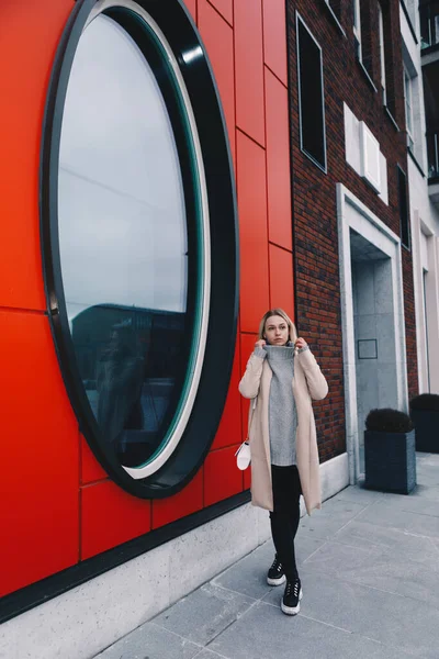 Long haired blonde woman wearing a sand coat with wool sweater, and black jeans, walking on a modern street around round window. Talking on the phone