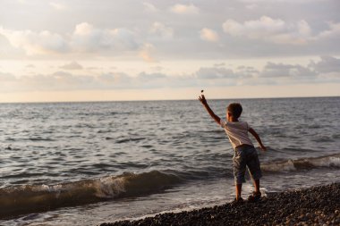 Çocuk gün batımında deniz kıyısına çakıl taşı atıyor.