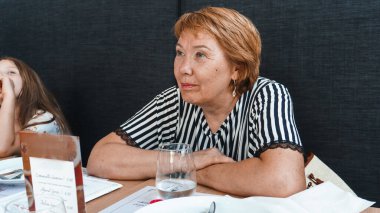 Masada oturan ve kameraya bakan yaşlı kadın. 