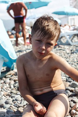 Çocuk sahilde oturuyor ve kameraya bakıyor.