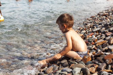Küçük çocuk sahilde oturuyor Denizde taşlarla 