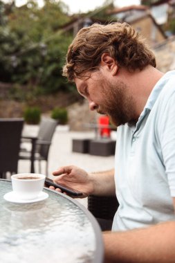 Kafede kahve ve telefonla oturan sakallı bir adam.