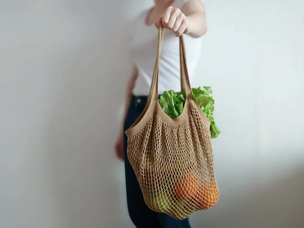 Knitted string bag with products oranges and greens. the shopper is beige. crochet eco bag in the hand of a girl in a white t-shirt and jeans.