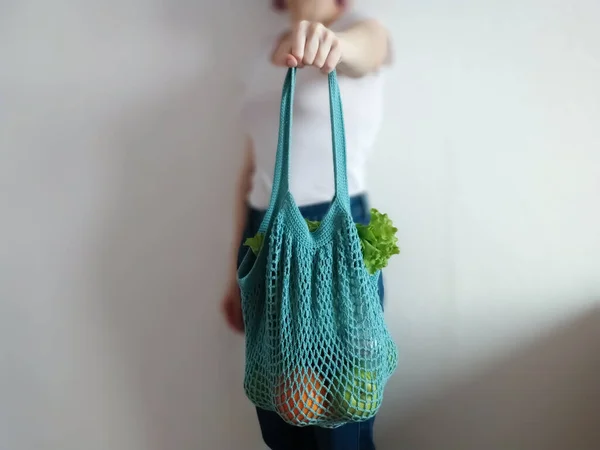 Knitted string bag with products oranges and greens. the shopper is blue. crochet eco bag in the hand of a girl in a white t-shirt and jeans.