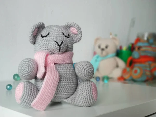 Crochet sleeping grey bear in pink scarf on background of other toys on light background. Knitted soft toy Teddy — Stock Photo, Image