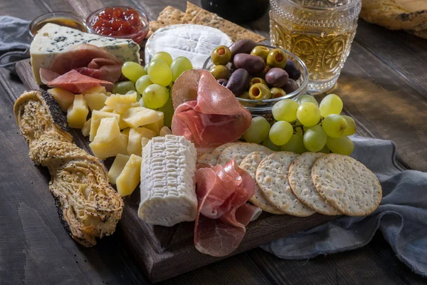 Plato de queso servido con uvas, mermelada, jamón, galletas saladas sobre un fondo de madera —  Fotos de Stock