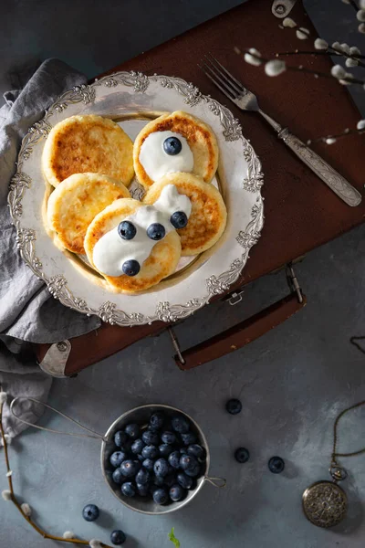 Cottage cheese pancakes on a vintage plate on dark background. Syrniki with fresh blueberries and sour cream. Pancakes with cottage cheese recipe. Top view, vertical. Russian cuisine