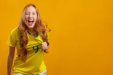 Brezilya taraftarı. Brezilyalı kızıl saçlı kadın fanatik futbolu, sarı arka planda futbol maçını kutluyor. Brezilya renkleri.