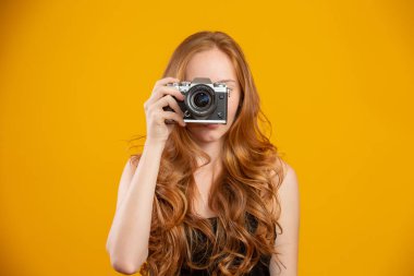 Siyah elbiseli, elinde antika fotoğraf makinesi olan ve sarı arka planda izole edilmiş fotoğraf çeken sevimli kızıl saçlı kadının fotoğrafı. Kopyalama alanı oluşturun. Eski model fotoğraf makinesini tut..