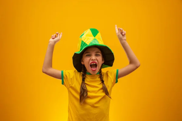 Football supporter, Brazil team. World Cup. Beautiful little girl cheering for her team on yellow background