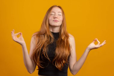 Kızıl saçlı beyaz kadın sarı arka planda duruyor rahatla ve gözleri kapalı parmaklarıyla meditasyon hareketi yaparken gülümse. Yoga konsepti.