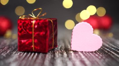 Altın düğümlü kırmızı hediye kutusu ve yakın plan pembe kalp. Arka planda parlak dekoratif toplar var. Noel ağacı ışıkları sarı yanıp sönüyor. Yeni yıl ve Noel havası. Gelecek yıl bayram hediyeleri ve aşkın beklentisi