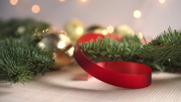 生生不息的云杉的枝条 装饰着新年的装饰品 背景是一个闪烁的花环 一只手把一个银色闪亮的球放在一个红色明亮的缎带旁边 圣诞前夕及新年前夕 — 图库视频影像