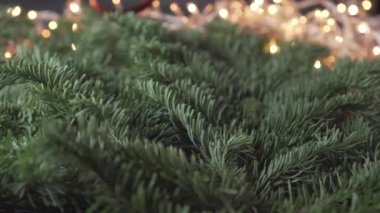 Bir adam, parlak kırmızı dekoratif kurdelelerden oluşan bir spirali yeni yıl ağacının yemyeşil dallarına, yanıp sönen sarı ışıklı bir çelengin arka planına serer. Noel ve Yeni Yıl dekorasyonları