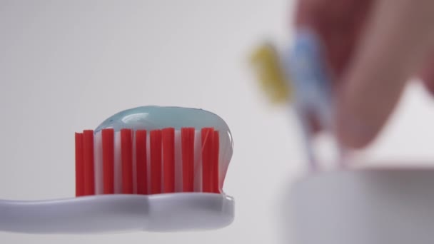 上に青歯磨き粉付きの赤い毛の白い歯ブラシ 背景には 手がガラスから青い歯ブラシを拾う 朝衛生上の手順 — ストック動画