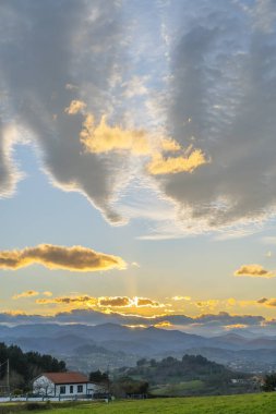 Ev ve çiftlik arazileri dağlarda gün batımı ve dağ zirvelerine karşı. Mavi gökyüzüne karşı turuncu arka plan ışığı olan sirus ve kümülüs bulutları. Güzel kırsal alan
