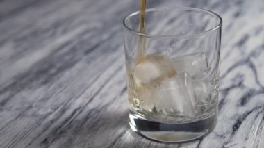 Golden whiskey is poured into a glass cup with ice cubes on an old wooden gray bar counter. Slow motion