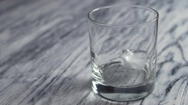 Frozen ice cubes fall into an empty glass for whiskey on an old wooden gray bar counter