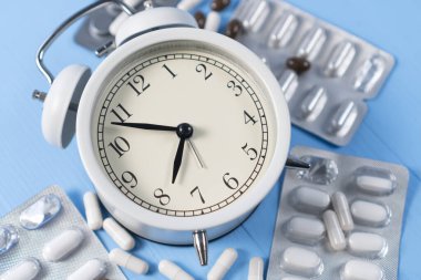 Açık mavi ahşap arka planda siyah okları olan alarm saati olan ilaç ve kapsüller yığını. Reçeteli ilaç alma düşüncesi