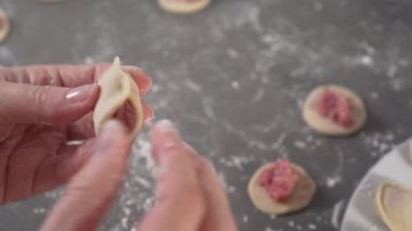 Kız elleri hamur ve etten ev yapımı ravioli heykeli yapıyor ve gri bir masa üzerinde içinde malzemeler ve un var. Beyaz bir tabakta yemek hazır.