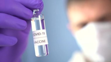 COVID-19 aşısı. Koruyucu eldivenler ve yüz maskesi takmış bir doktor elinde mavi arka planda koronavirüs ilacı olan bir ampul tutuyor. Yeni bir virüsün enjeksiyonunu ara ve uygula