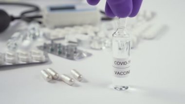 Coronavirus aşısı. Mavi koruyucu eldivenli bir el, COVID-19 ilacının bulunduğu ampulü tıbbi ekipman, tablet ve kapsüllerle dolu bir masaya yerleştirir.