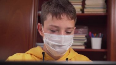 Dizüstü bilgisayarın önünde koruyucu maske takan bir genç koronavirüsten karantinaya alınırken internetten öğreniyor. Kitaplık ve okul malzemeleri olan bir oda. Antiviral karantina sırasında internet üzerinden ödev yapmak