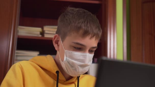 自宅のノートパソコンの前で顔のマスクで10代の若者の距離学習 コロナウイルスからの隔離 インターネット上で宿題をする — ストック動画