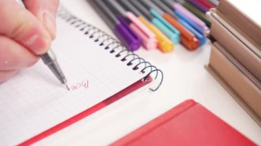 Genç adam beyaz bir masanın üzerine parlak okul malzemeleriyle birlikte bir defter yazıyor. Çok renkli keçeli kalemler ve kitaplar. Yakın plan.