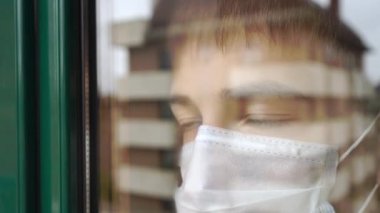 Karantinada genç bir adam var. Tıbbi maske, üzgün gözlerle pencereden dışarı bakar. Şehir binaları yansımada. Virüs koruması için kendini izolasyon