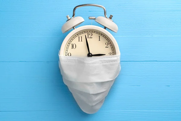 Beyaz tıbbi maskeli çalar saat. Mavi, ahşap, parlak bir arka planda. Coronavirüs 'ten ekonomik ve bankacılık krizi kavramı.