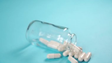 Mavi arka planda beyaz tıbbi kapsüller serpiştirilmiş bir ilaç şişesini kapat. COVID-19 tedavisi. Coronavirus aşısı konsepti. Kamera yavaşça aşağı iniyor.