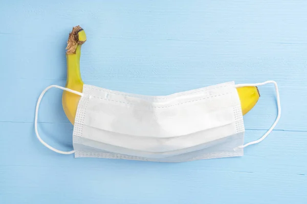 Yellow ripe banana covered with a white medical protective mask on a wooden light blue background. Close-up. The concept of protected sex and contraception. Strengthening immunity for the treatment of coronavirus