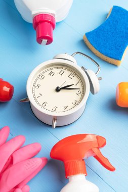 Sünger, pembe koruyucu eldiven ve alarm saati olan çok renkli şişelerdeki deterjanlar açık mavi ahşap arka planda. Ödev konsepti