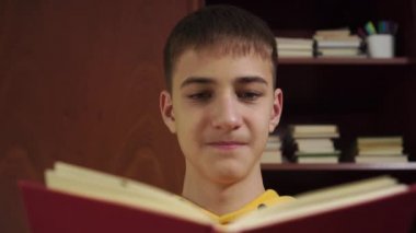 Genç öğrenci bir kitap okuyor ve arka plandaki ahşap kitaplıkta ders kitapları ve okul gereçleri ile gülüyor. Sarı bir kazakla. Yakın plan. İzolasyonda çevrimiçi uzaklık öğrenimi
