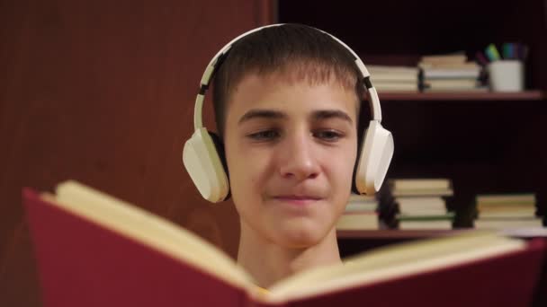 教科書や学校用品のある本棚を背景に 白いワイヤレスヘッドフォンで本を読む青年 接近中だ オンライン学習へコロナウイルスの隔離 — ストック動画
