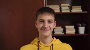 Genç bir öğrenci, arka plandaki kitaplık ve okul gereçleri ile beyaz bir kupadan gülüyor ve içiyor. Yakın plan. Korornavirüs karantinasında günaydın.