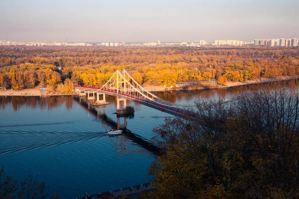 Brug voor wandelingen over de rivier tegen de achtergrond van herfstbomen en een drijvende boot op de rivier — Stockfoto