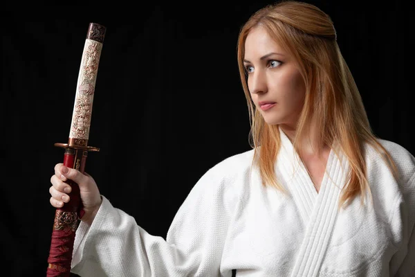 Vacker flicka med blont hår i vit kimono håller en japansk katana svärd mot en svart bakgrund — Stockfoto