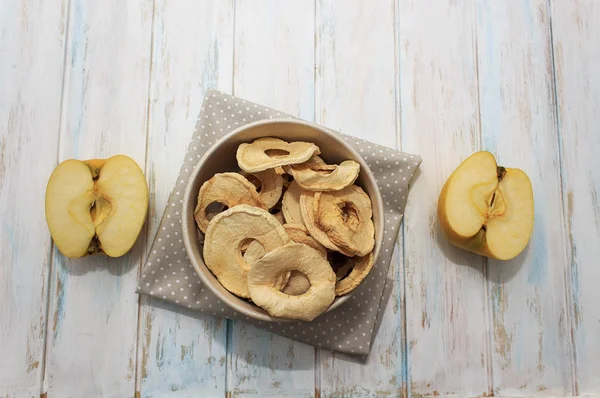 Dried apple chips in a bowl on a white wooden background and towel. Diet snack with copy space. Top view